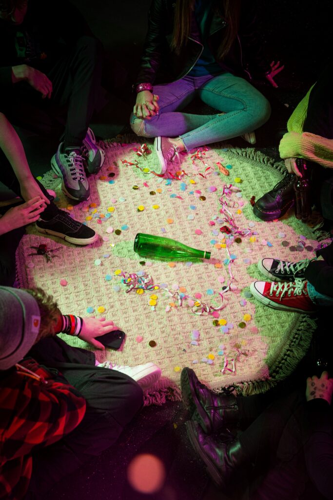 A group of youngsters sit in a circle around a green bottle with their feet protruding into the circle. They are wearing an eclectic collection of shoes - baseball boots, doc martens and trainers. There's confetti and streamers on the carpet, alongside a set of car keys and some money. The image is tinged with pink and green hues reminiscent of party lights.