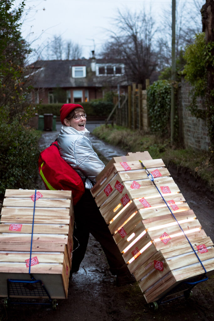 Performer Fiona Manson, wearing a silver puffer jacket and carrying a red courier delivery pack, is walking down a driveway with two stacks of crates on trolleys. She is smiling and looking backwards at the camera.