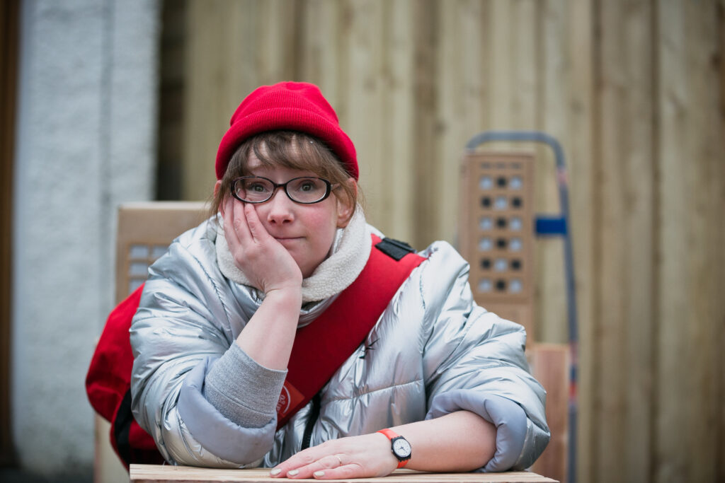 Performer Fiona Manson in a silver puffer jacket and red hat, rests her elbow on a stack of boxes she's delivering.