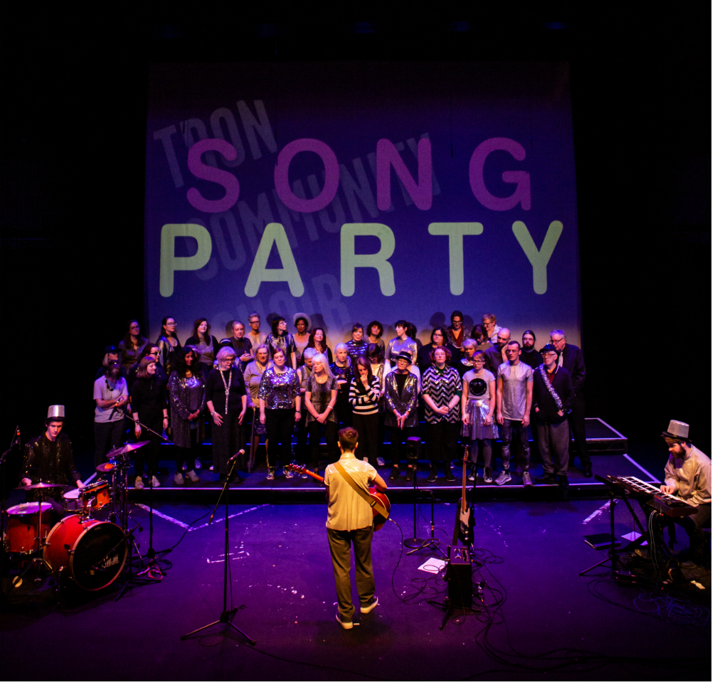 Tron Community Choir members, all wearing silver clothing, performing on the main stage in front of a purple backdrop that says Song Party. The choir impresarios are in the foreground leading the singing, from left to right, playing drums, guitar and keyboards.