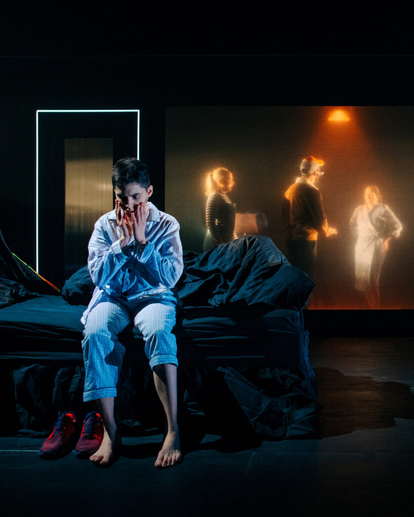 A man in blue pyjamas sits on the edge of an unmade bed. He has his hands to his face in shock. Behind him through a hazily-lit window are three people seemingly going about their daily tasks.