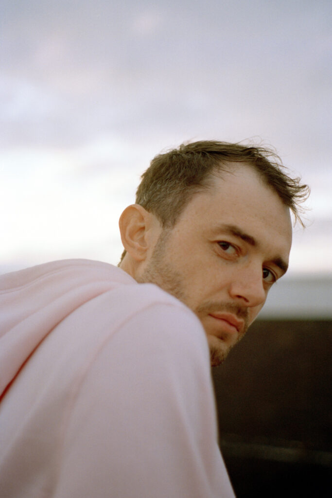 A dark-haired, bearded man in a pink hooded top stares backward over his shoulder to the camera. He's photographed outside against a grey sky.