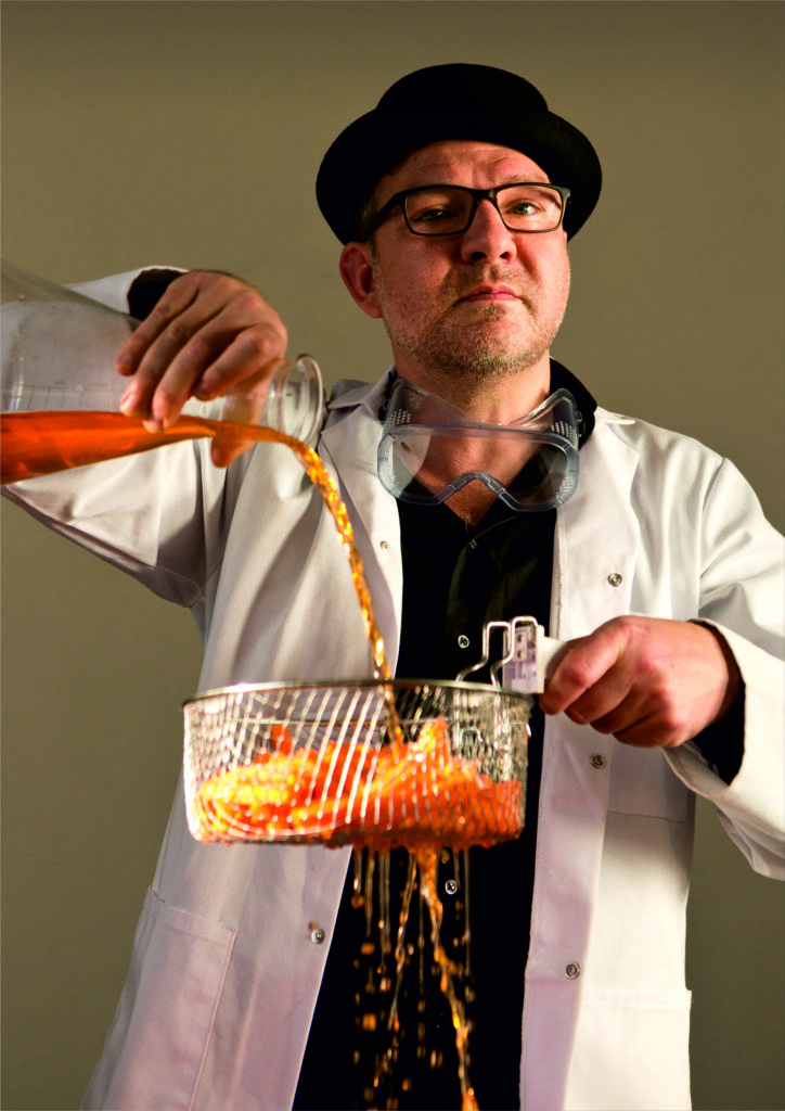 A man in a white overall and black pork-pie hat pours vinegar from a glass vessel over metal basket of chips.