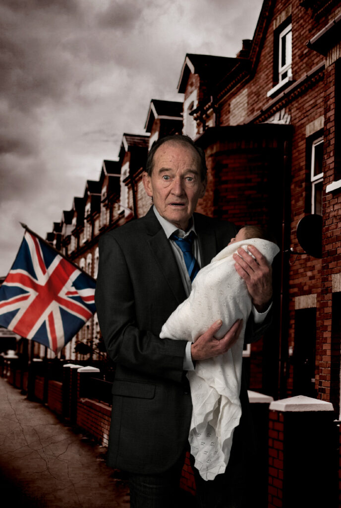 Cyprus Avenue promotional image. A bewildered-looking older man stands in a street of red-bricked terraced houses. He holds a small baby wrapped in a white blanket. There's a Union Jack flag flying from one of the houses behind him. The sky is has dark storm clouds forming.