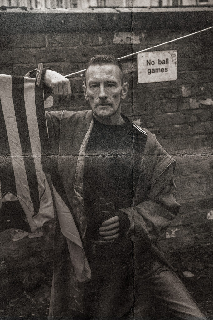 Moorcroft promotional image. An older man in a parka and holding a pint stares directly into the camera. He is standing against a brick wall with a sign saying No ball games. He has his arm draped over a washing line. A striped football top blows in the wind.