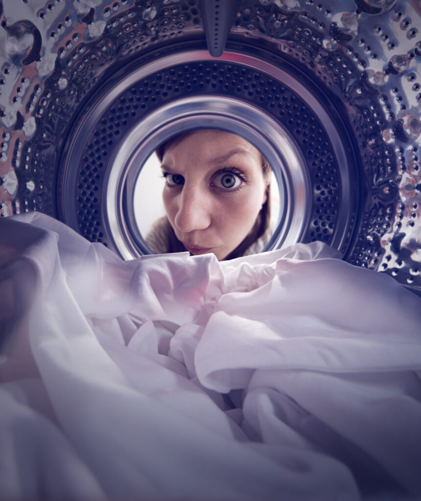 Spin! promotional image. Taken from inside the drum of a washing machine, a dark-haired woman peers through the circular door at the washing in the drum.