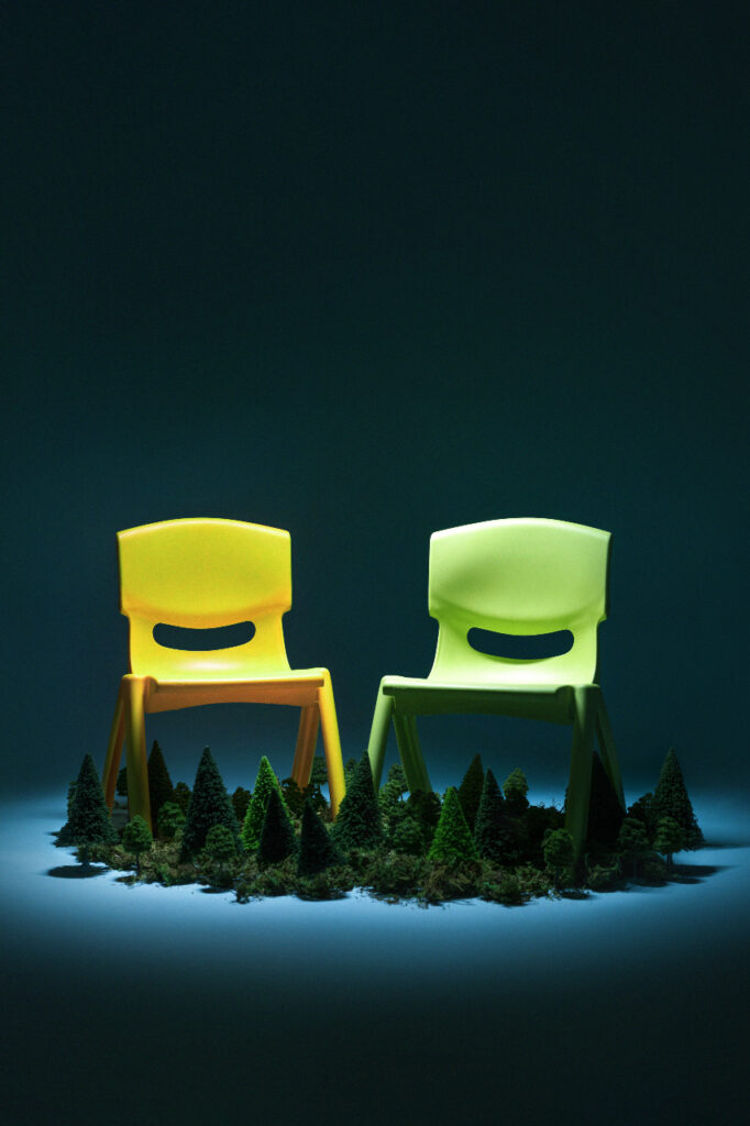 Wolfie promotional image. Two small plastic chairs, one yellow and one green sit amidst a forest of tiny green fir trees. The cut outs on the back of the chair look like innocent child-like smiles. The dark background and moody lighting give the chairs a sense of malevolance.