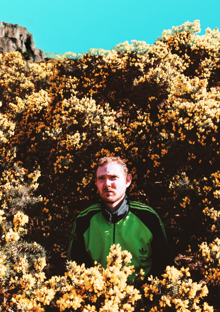 A young man with scruffy hair and beard and wearing a bright green and black tracksuit top is surrounded by yellow gorse bushes in full flower.