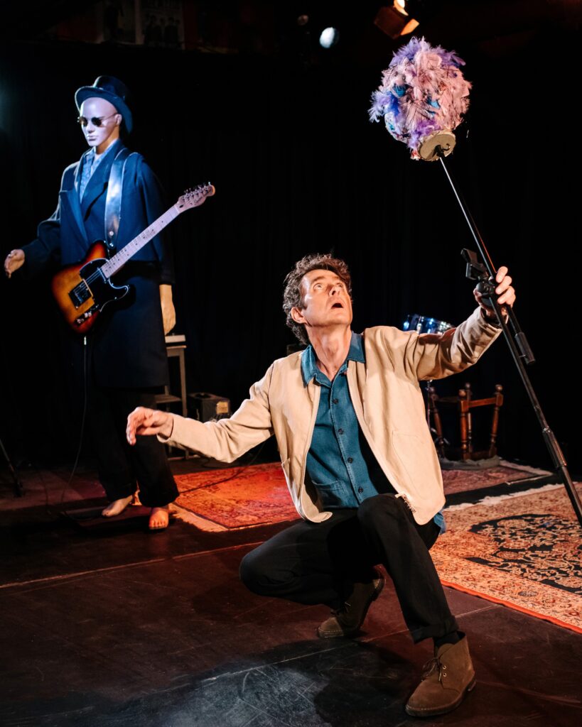 A man in a denim shirt, light suede jacket, black trousers and brown suede desert books crouches down on a stage with a microphone stand in his hand. He is looking up to a pink and purple feathered object attached to the mic stand. Behind him a shop mannequin dressed in a suit, sunglasses and a hat 'plays' guitar.