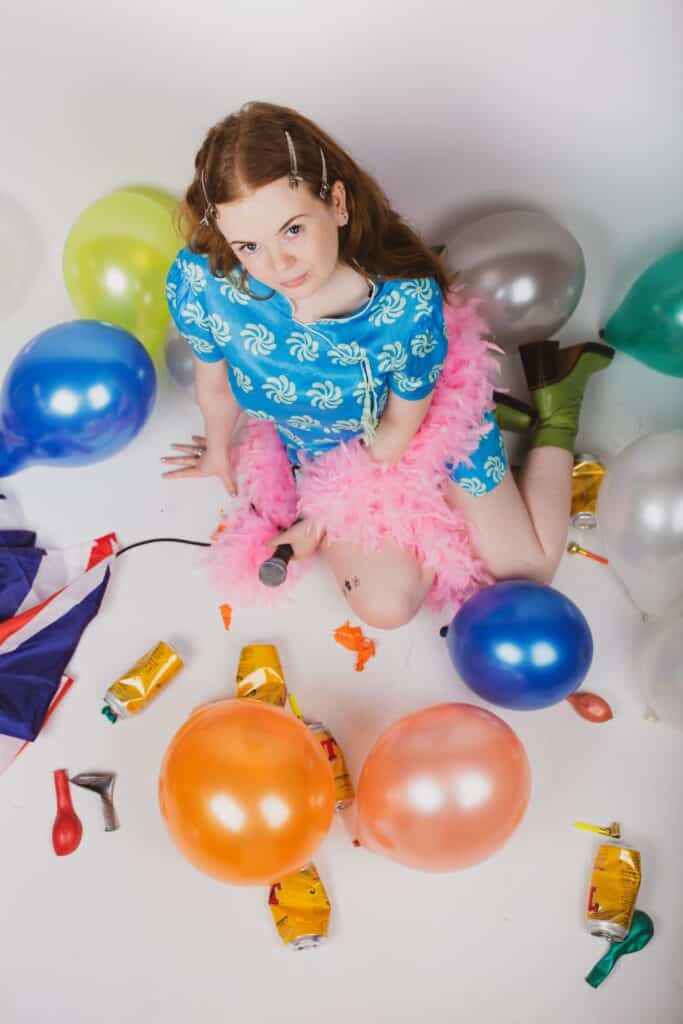 A young woman in a turquoise short dress, green heeled boots and pink feather boa is photographed from above. She looks up to the camera with as impassive stare. She's surrounded by balloons, beer cans, a union jack flag and party blowers. She is holding a microphone in her left hand.