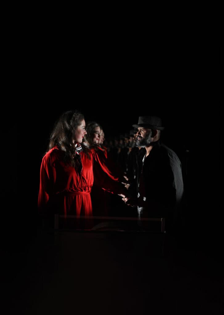 A man in a black hat and coat holds the hand of a long dark-haired woman in a red dress. The are photographed in front of a mirror, so are reflected multiple times in the background. The woman smiles and looks lovingly at the man, whilst he looks serious and sad.