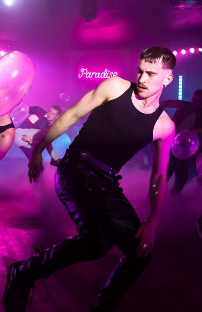 A young, muscular man in a black vest, leather trousers and heavy black boots dances in a neon pink-lit nightclub. The word Paradise in pink neon lights up the background. Several other men appear in the background doing the same angular dance move.