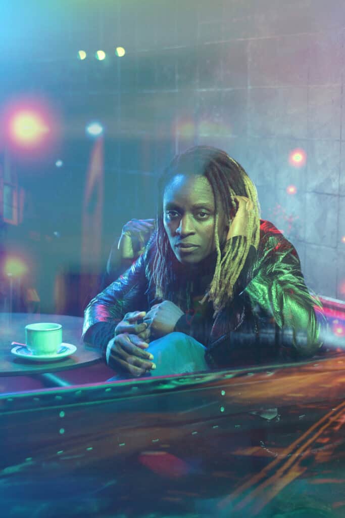 A black woman with long dreadlocked hair and wearing a black leather coat and jeans stares out of the window of a cafe. There's a coffee cup on the table in front of her and the lighting suggests it is night time. The image is distorted in a way that shows her hands in multiple different positions, reflecting her different states of mind.