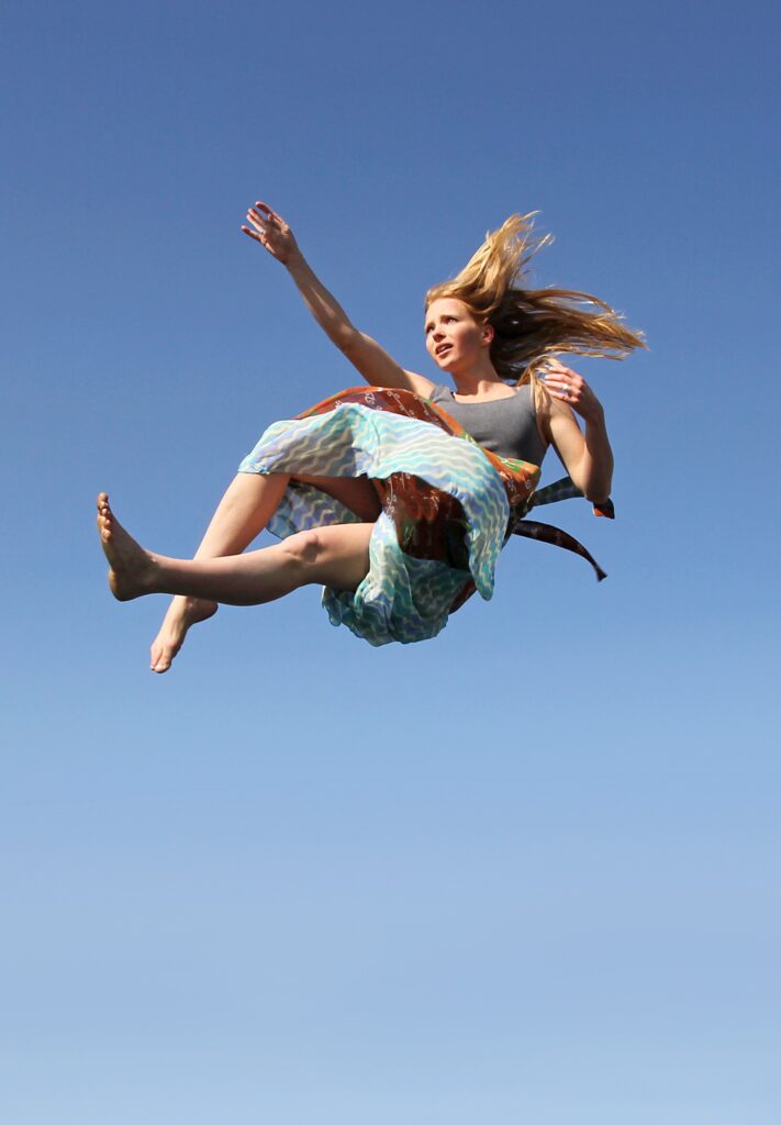 A woman is photographed against a deep blue sky. She appears to be floating in the air. Her hand and leg are outstretched, her long blond hair and clothes blow in the wind. She has a concerned expression.