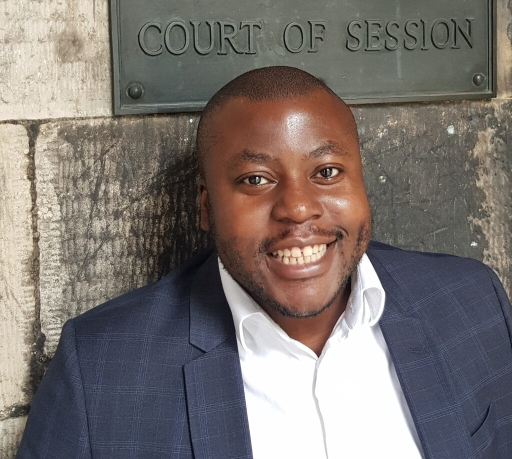 A black man wearing a white shirt and blue suit jacket looks directly to camera, smiling. He stands in from of a stone wall. Above his head is a plaque that reads Court of Session.