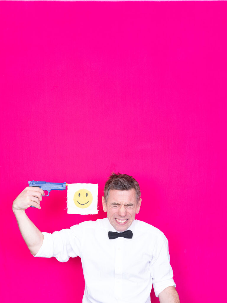 A man in a white shirt and black bow-tie is photographed against a bright pink background. He holds a toy gun to his head which has a white flag with a smiley emoji printed on it coming out of the barrell. His face is screwed up into a grimace.
