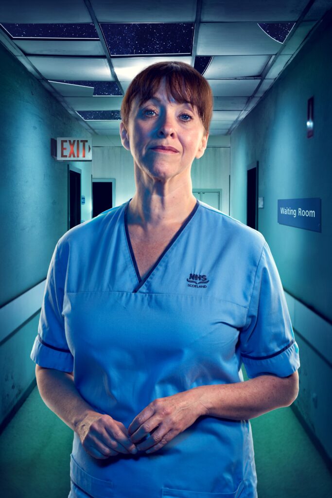 A dark haired woman wearing a blue nurses uniform stands in a hospital corridor. The tiles on the ceiling above her are missing or broken, exposing a dark starry night sky.