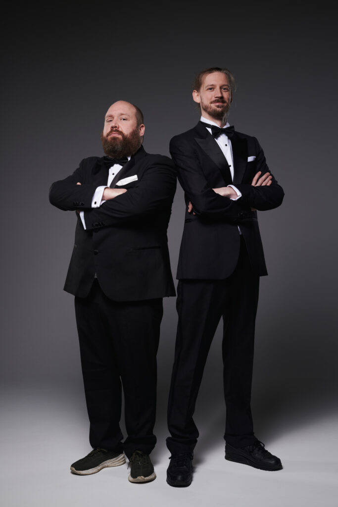 Two bearded men in black tuxedo suits, white shirts and bowties look straight to camera. Both have their arms folded and are wearing scruffy trainers. The man on the left has a stern expression whilst the man on the right has a wry smile.