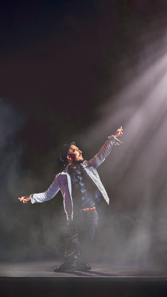 A beared male dancer wearing a bowler hat, light denim jacket, dark jeans with a brown belt and black trainers adopts a balletic pose - with right foot pointed and arms outstretched. He's photographed on a dark stage surrounded by haze and looks off to the right.