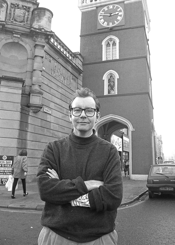 A black and white image of Michael Boyd, in a dark jumper and with his arms folded outside the Tron Theatre - with the Tron steeple in the background.